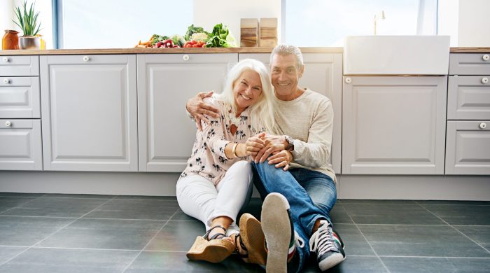 Happy mature attractive woman and handsome man embracing while sitting on kitchen floor. Large cabinets surround them.